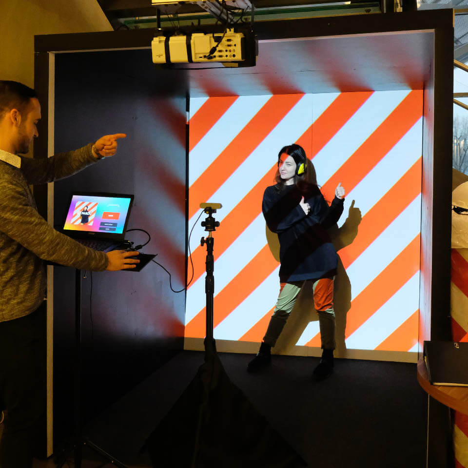 Lady in black outfit is standing inside a 2x2x2 metres black cube. On the back wall of the cube thick orange an white diagonal lines are projected using special projector. Camera on a tripod is recording her moves. This lady is posing to make a video in our projection cube photo booth