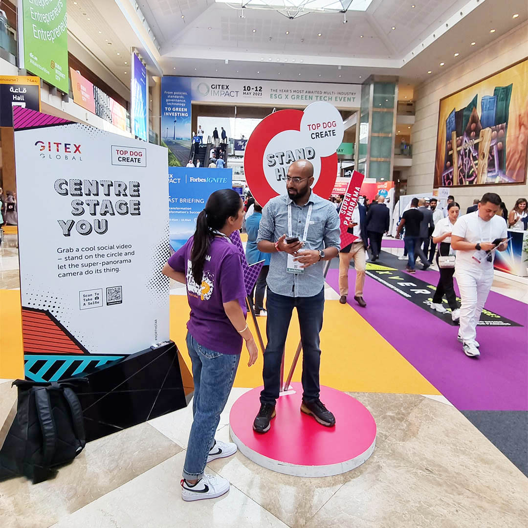 Man is making a video in Super Zoom Selfie Photobooth on an exhibition stand. Assistant is explaining to him how to do it. 